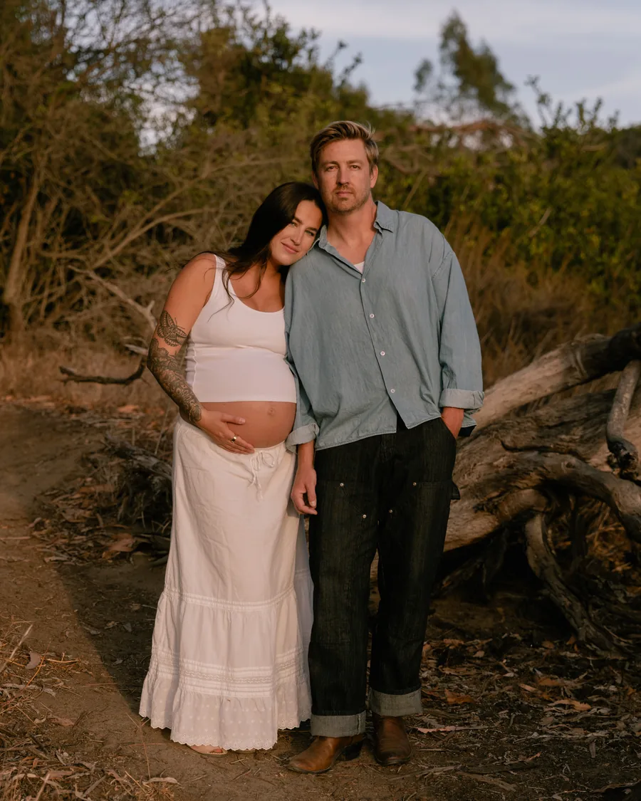 Expecting couple standing together on a dirt path at golden hour, her hand resting on the bump