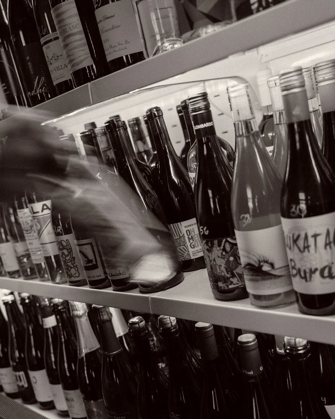 Black and white close-up of natural wine bottles on the shelves, a hand reaching in mid-motion