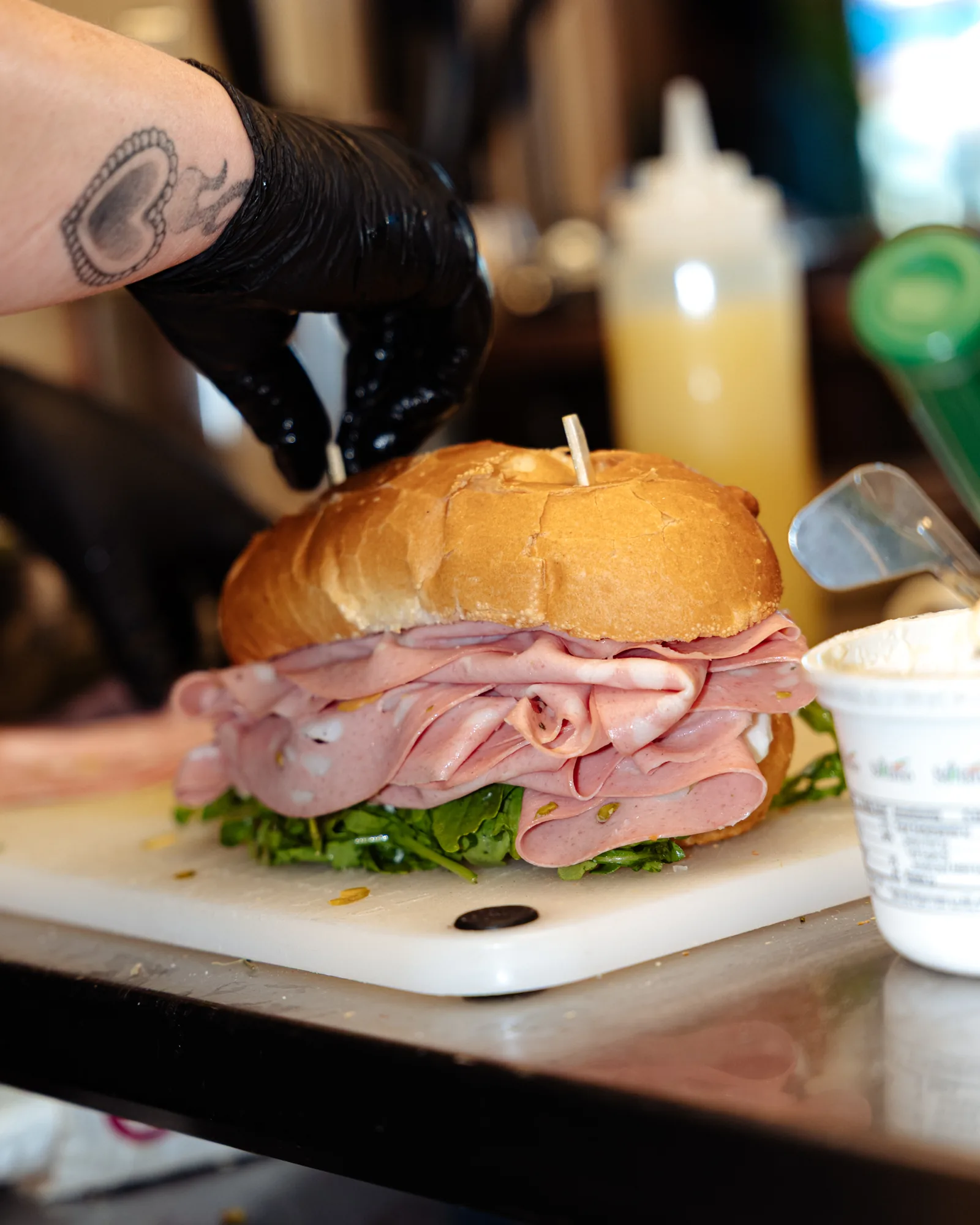 A gloved hand building a mortadella sandwich on a cutting board