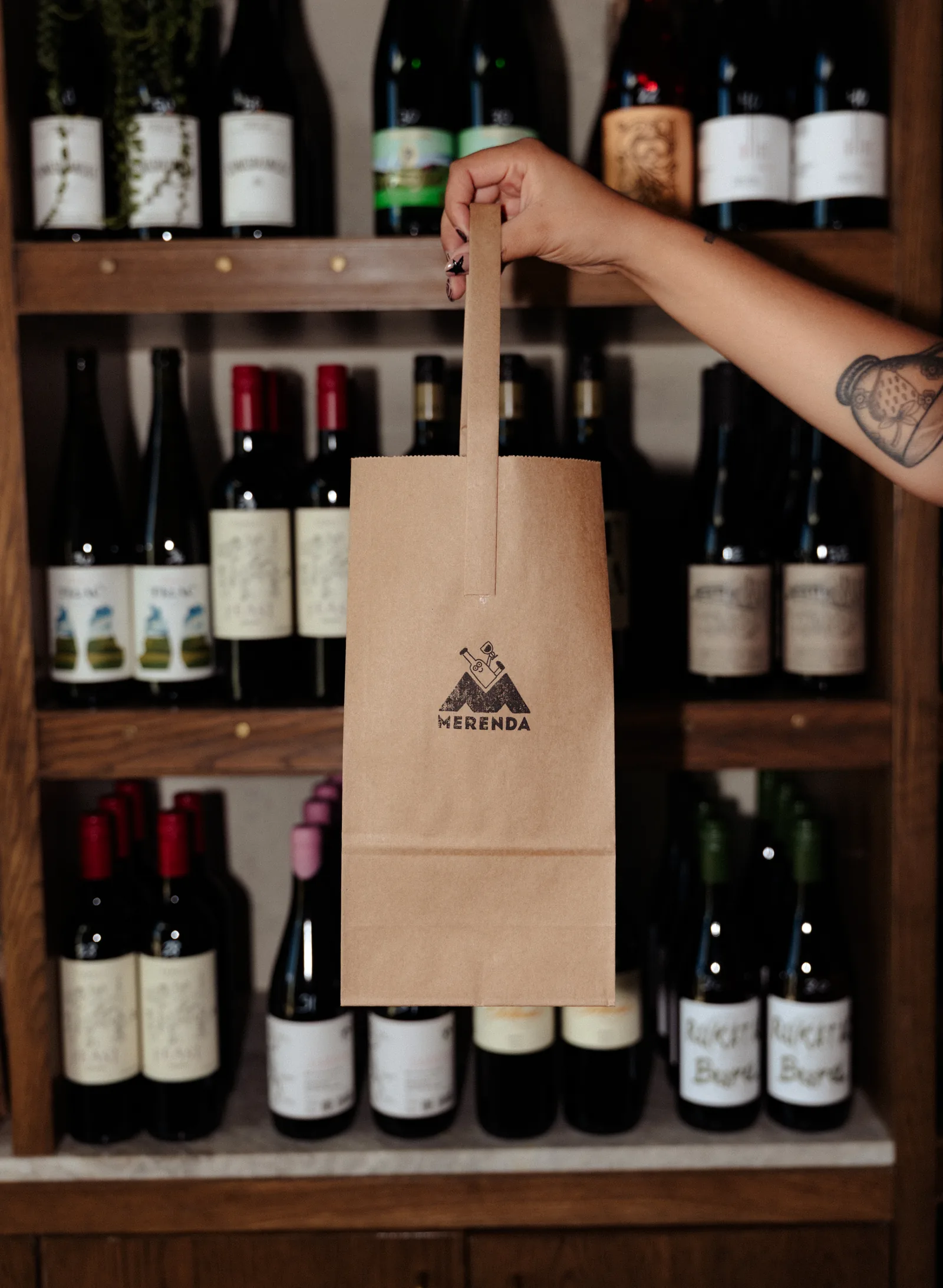 A hand holding a Merenda paper wine bag in front of shelves of bottles