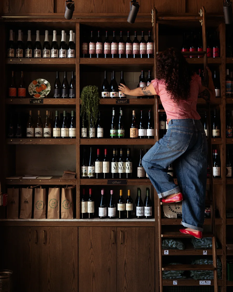 A woman on a rolling ladder reaching for a bottle high on Merenda's wine wall