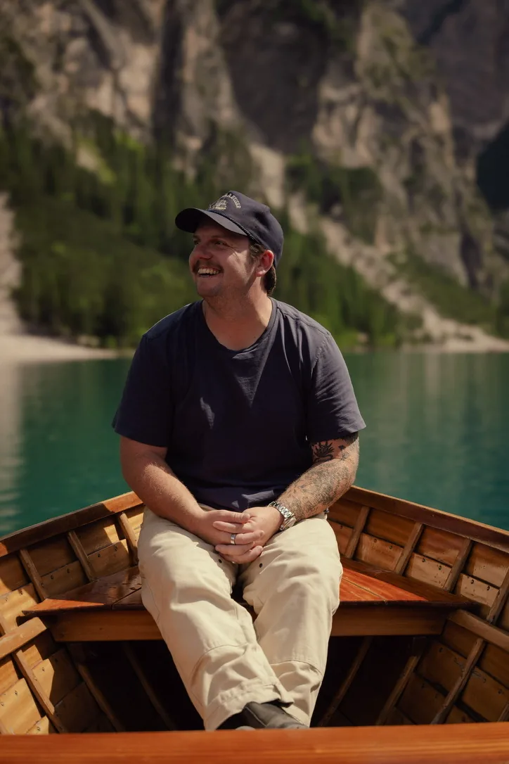 Michael Easterling smiling on a wooden boat with mountains and a turquoise lake in the background