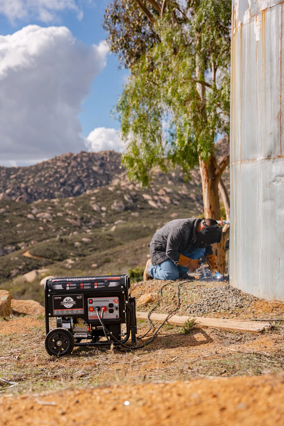Tomahawk welder generator on a hillside, welder crouched at a steel tank