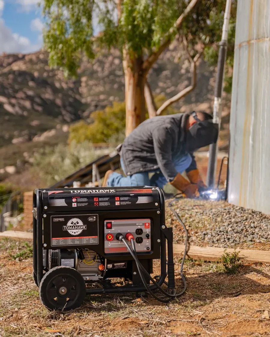 Tomahawk TWG210A welder generator running on a hillside job site, welder in a full-face helmet working on a metal structure behind it