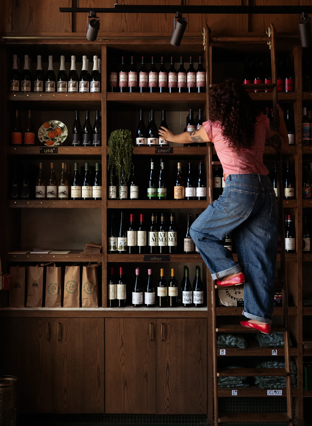 A woman on a rolling ladder reaching for a bottle high on the wine wall