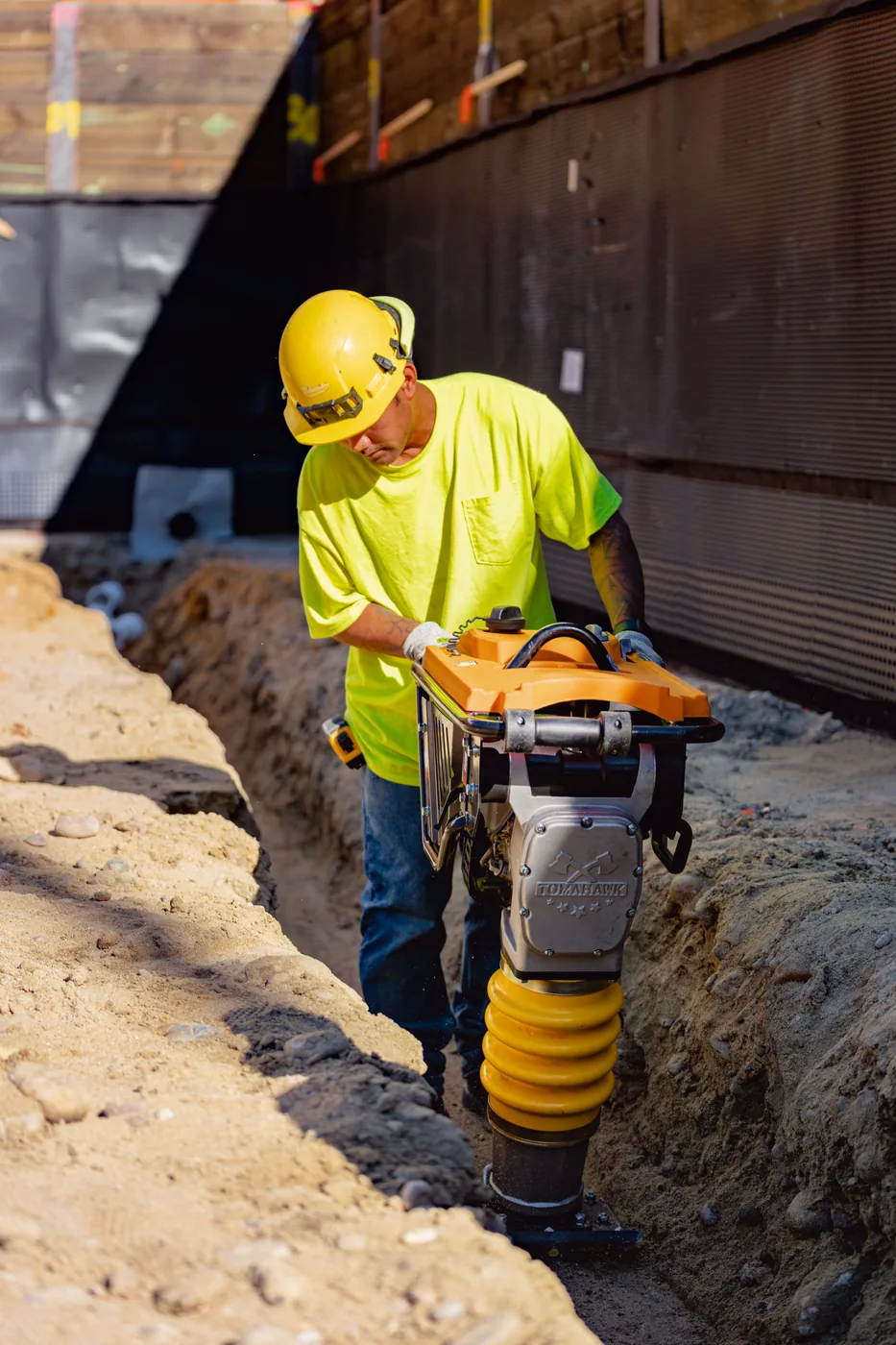 Worker lowering a rammer compactor into a narrow trench
